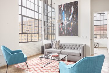 Modern loft living room with gray couch, blue accent chairs, mirrored coffee table, large metal floor lamp, orange and white rug, large industrial windows.